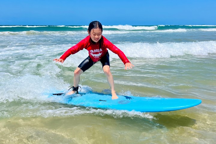 Child in red shirt surfing on blue board in shallow ocean waves.