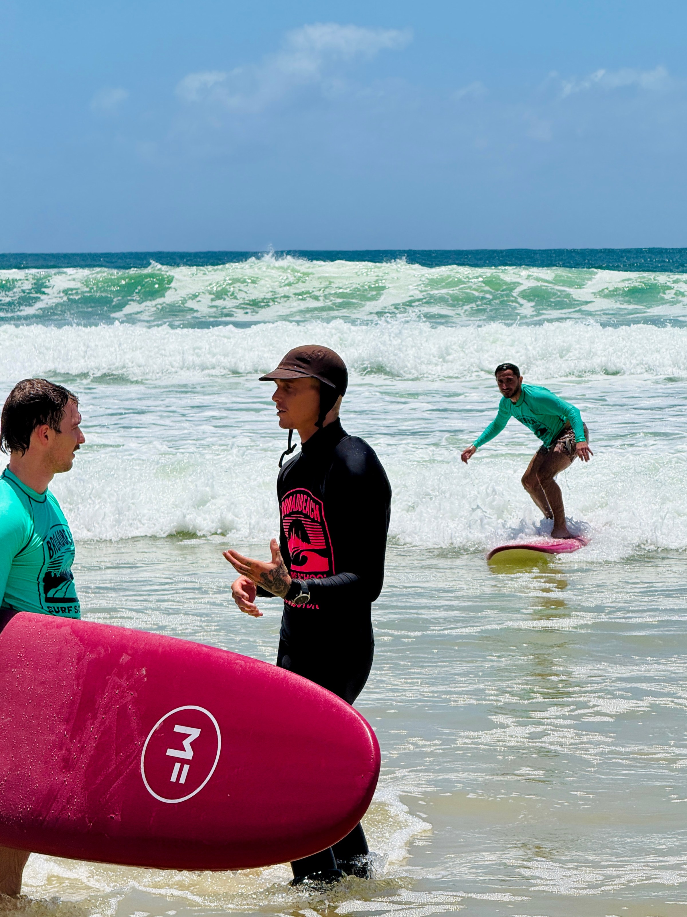 Three men in wetsuits at the beach; two with surfboards, one surfing.