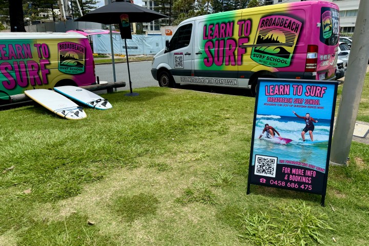 Surf school van and sign on grass with surfboards, promoting learn to surf lessons.