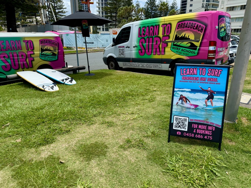 Surf school van and sign on grass with surfboards, promoting learn to surf lessons.