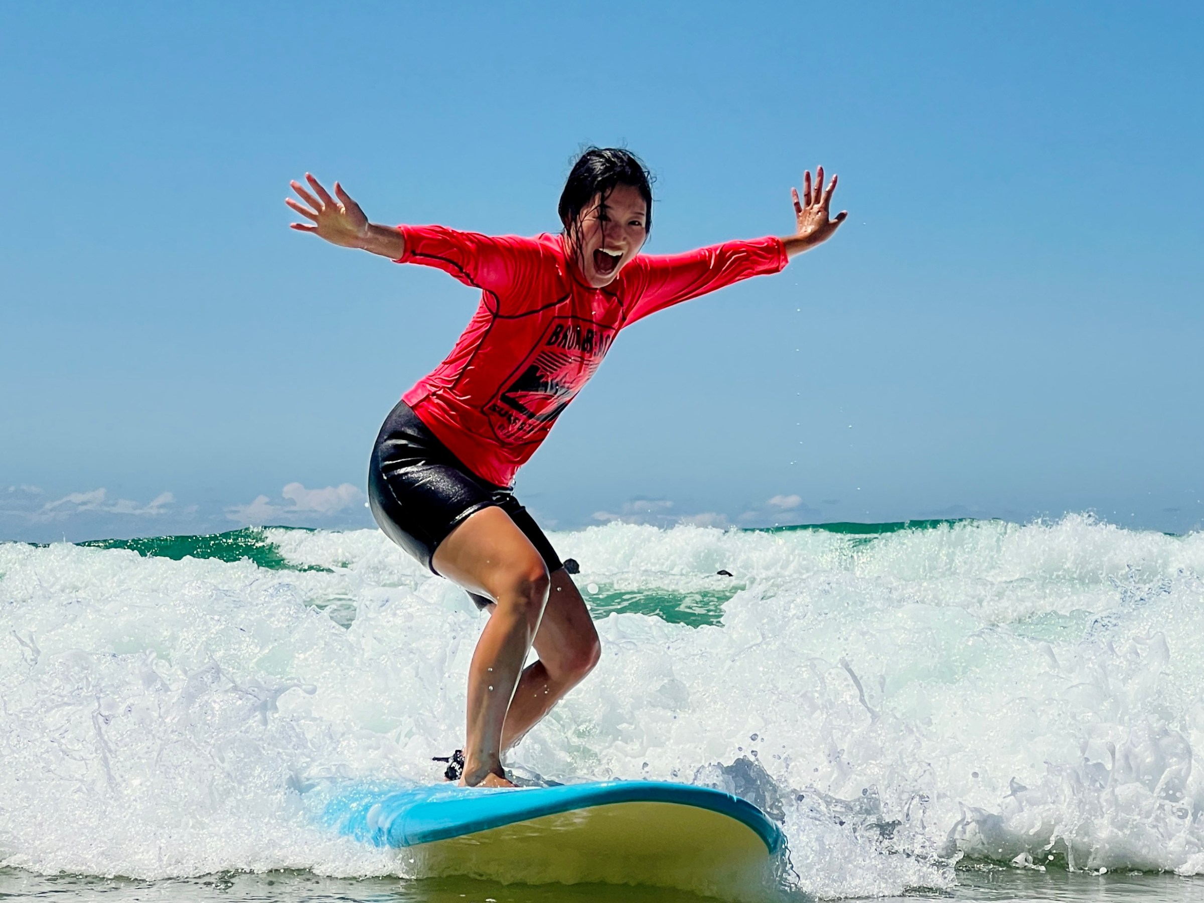 Person surfing on a blue board with arms outstretched, wearing a red shirt and black shorts, on a sunny day.