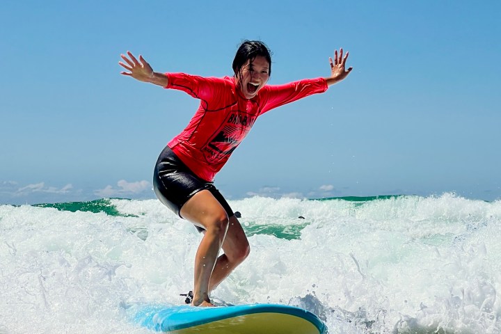 Person surfing on a blue board with arms outstretched, wearing a red shirt and black shorts, on a sunny day.