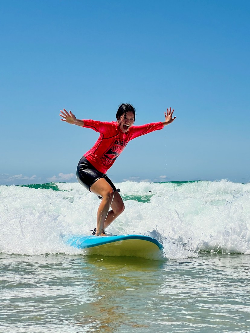 Person surfing on a blue board with arms outstretched, wearing a red shirt and black shorts, on a sunny day.