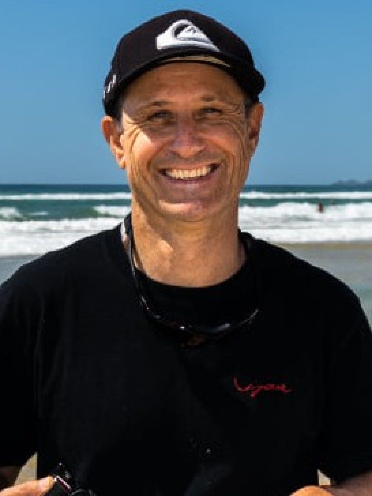 Smiling person wearing a black cap and shirt, standing on a beach.