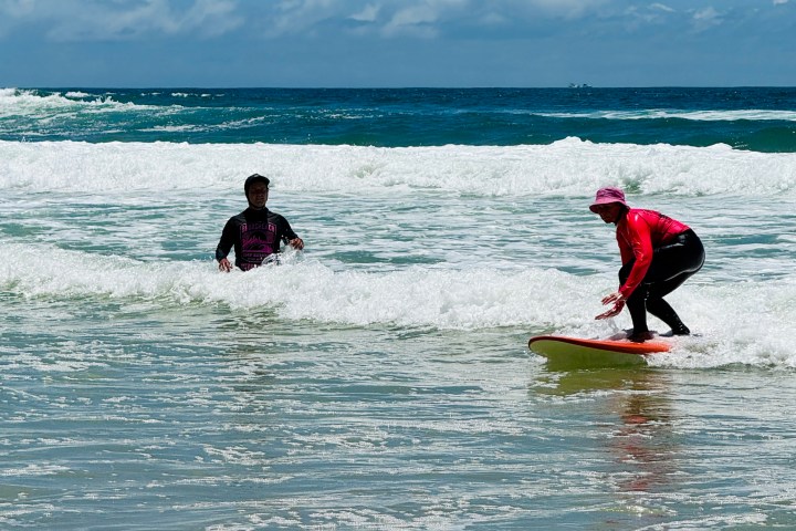 Two people in the ocean, one surfing on waves, under a clear blue sky.