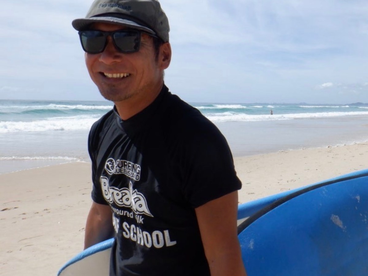 Person in sunglasses and cap holds a blue surfboard on a sunny beach.