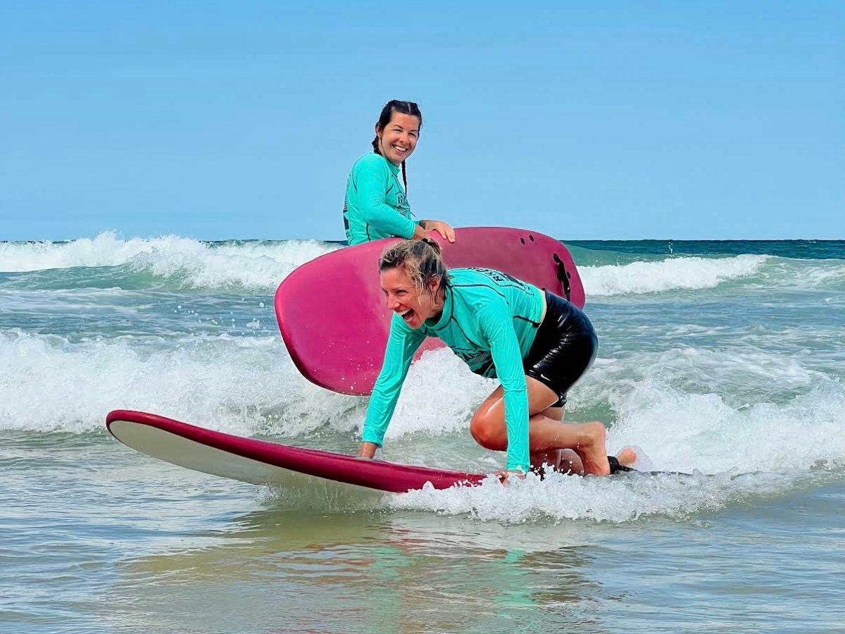 Two people surfing with pink boards and wearing teal tops on a sunny beach.
