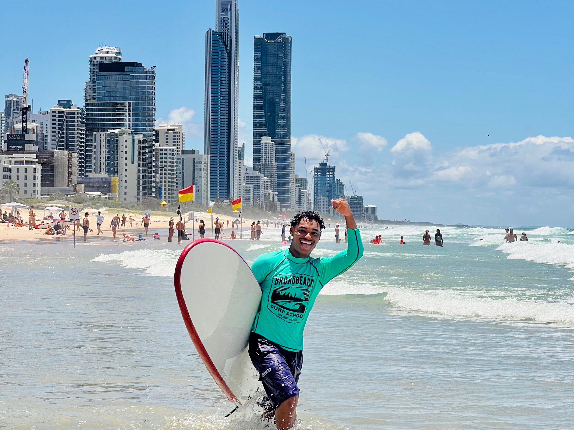 Man smiling with surfboard on sunny beach, city skyline behind.