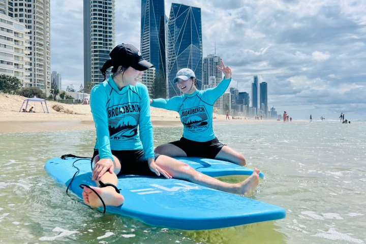 Two surfers sitting on boards near skyscrapers on a sunny beach.
