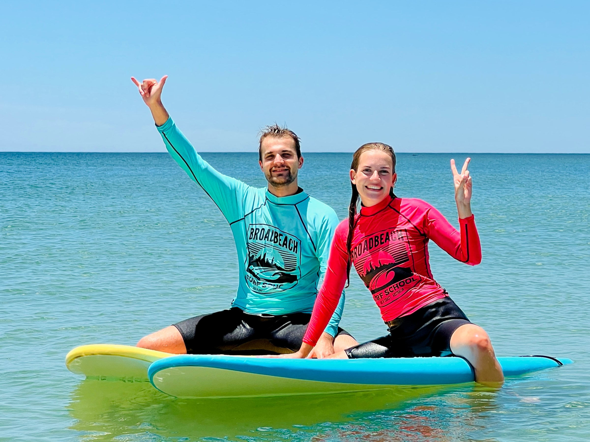Two people on surfboards in the ocean, smiling and gesturing peace signs.